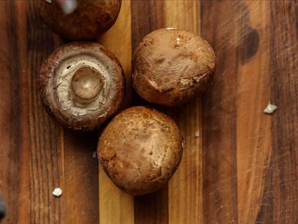 Four whole brown mushrooms lie on a striped wooden cutting board, with one mushroom showing its stem and gills.